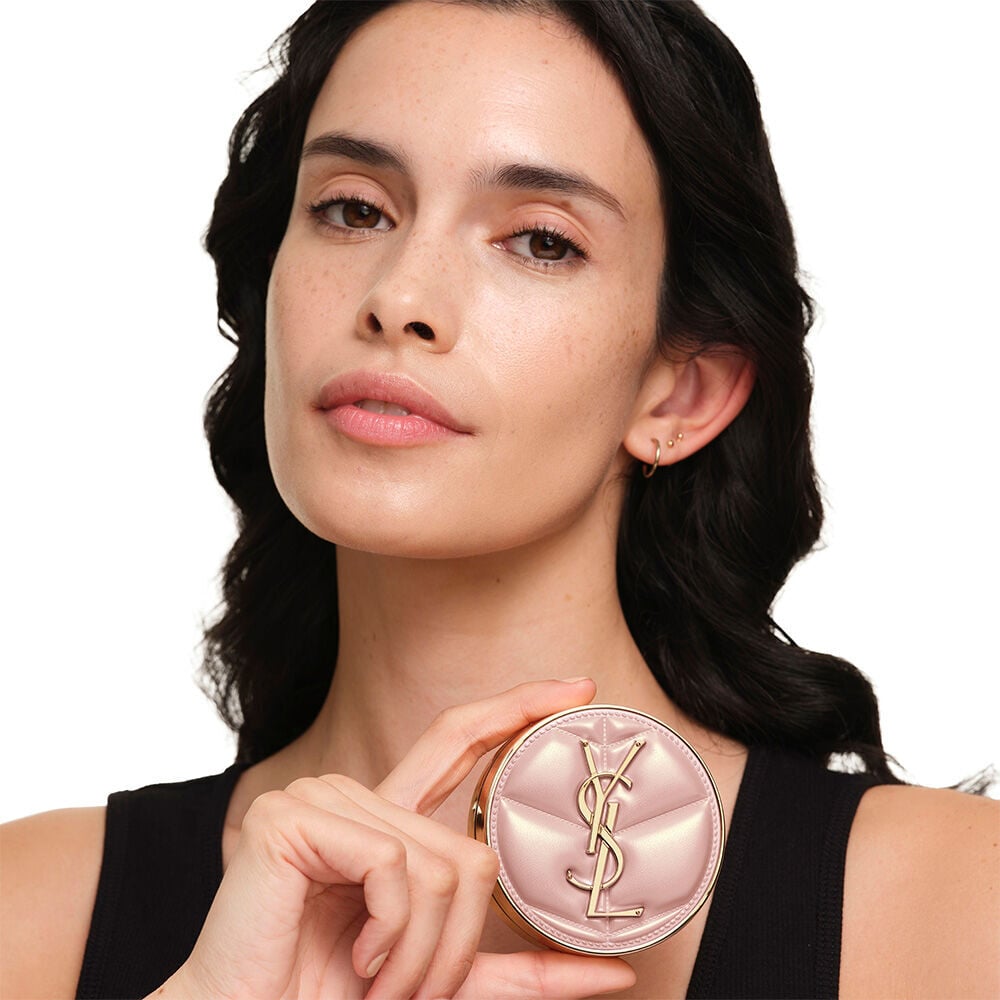 Model with a smooth, radiant complexion holding the pink YSL SKIN AFFAIR CUSHION FOUNDATION to showcase a naturally glowing, flawless skin finish.