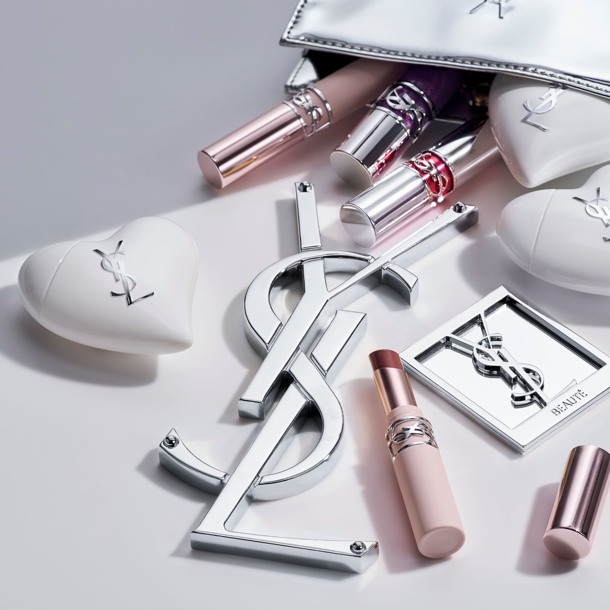 YSL makeup and Care Crush products spilling from a silver pouch around a silver Cassandre logo. Silver & white lifestyle.