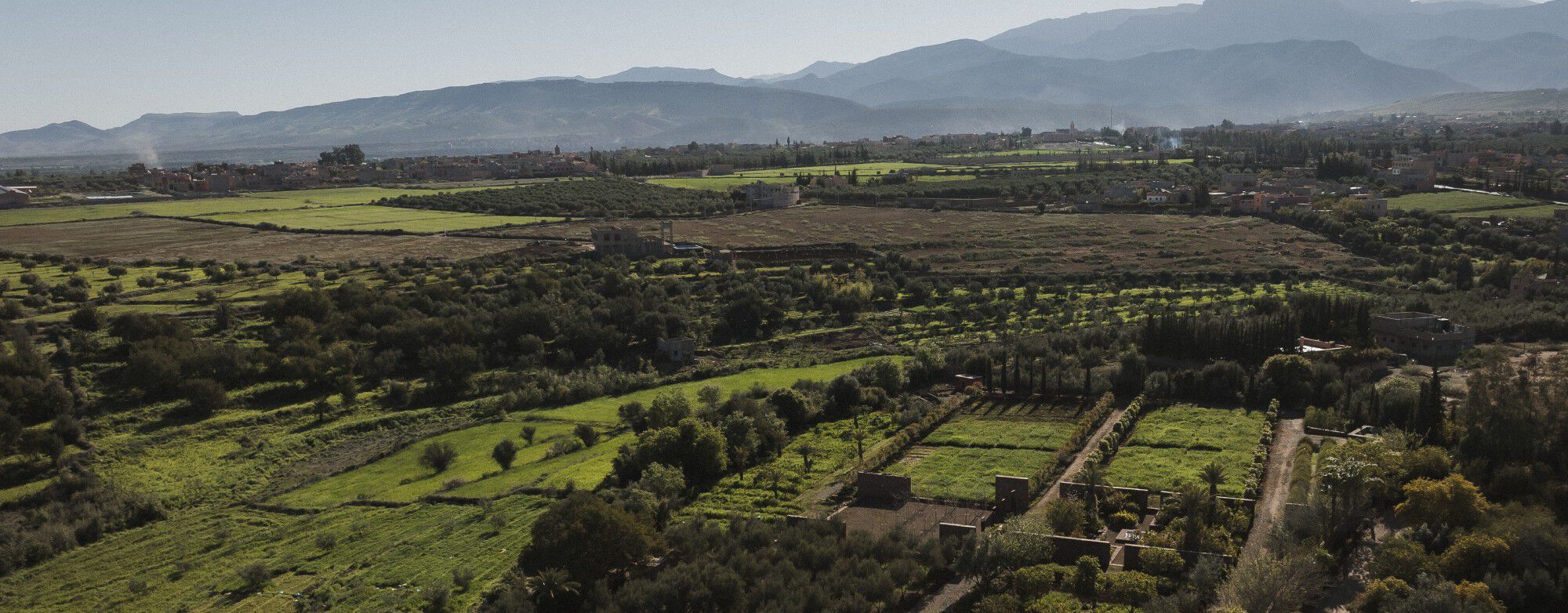 YSL pour l'avenir : Vue aérienne des jardins communautaires d'Ourika, champs verdoyants, arbres et montagnes sous un ciel dégagé.