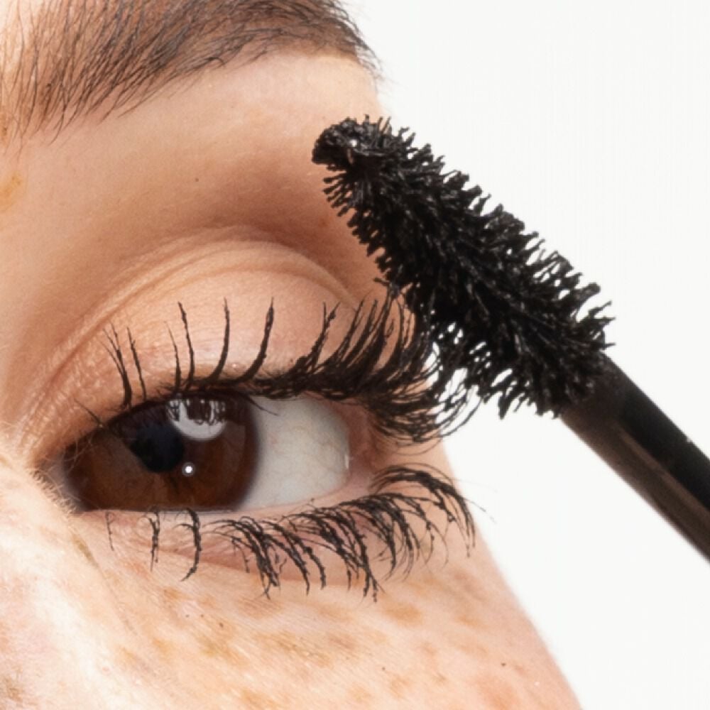 Close-up of a brown eye with long, voluminous lashes, as the YSL Lash Clash Mascara brush applies a rich, black formula.