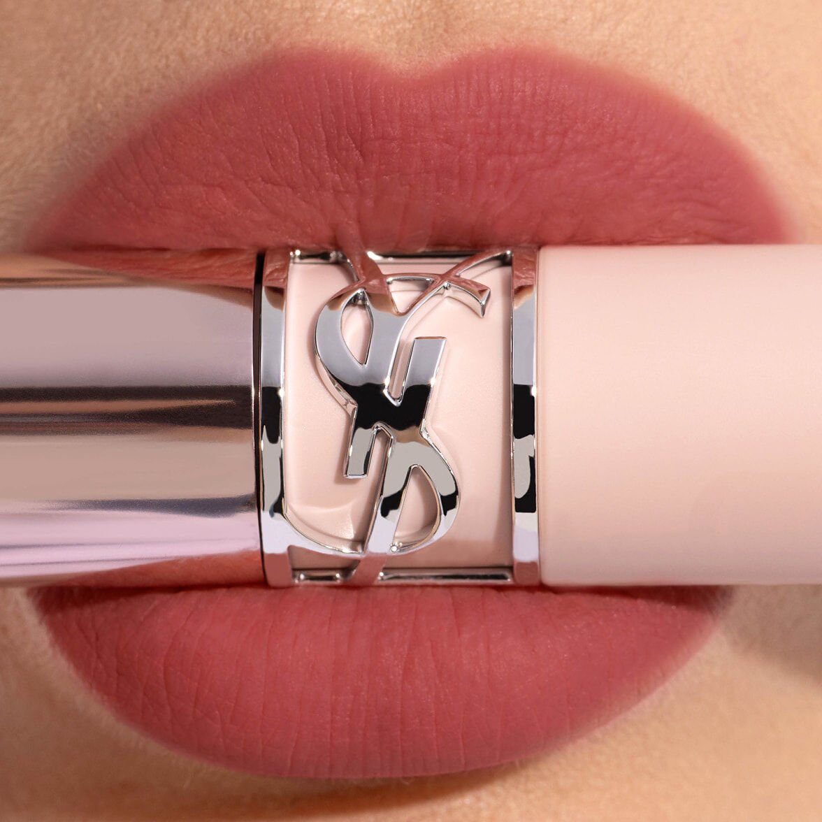 Gros plan sur des lèvres portant le YSL LOVENUDE LIP BLUSHER en teinte 4, avec un tube de rouge à lèvres YSL rose pâle et une bague Cassandre argentée..