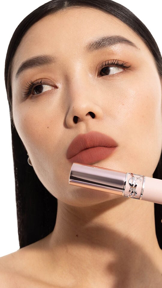 Le mannequin Mei J portant le YSL LOVENUDE Lip Blusher en teinte 3, tenant le produit près de ses lèvres nude rosées.