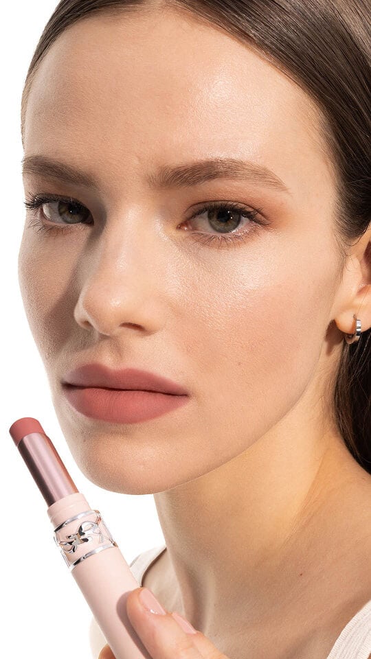Le mannequin Jade portant le YSL LOVENUDE Lip Blusher en teinte 2, tenant le produit près de ses lèvres nude rosées.