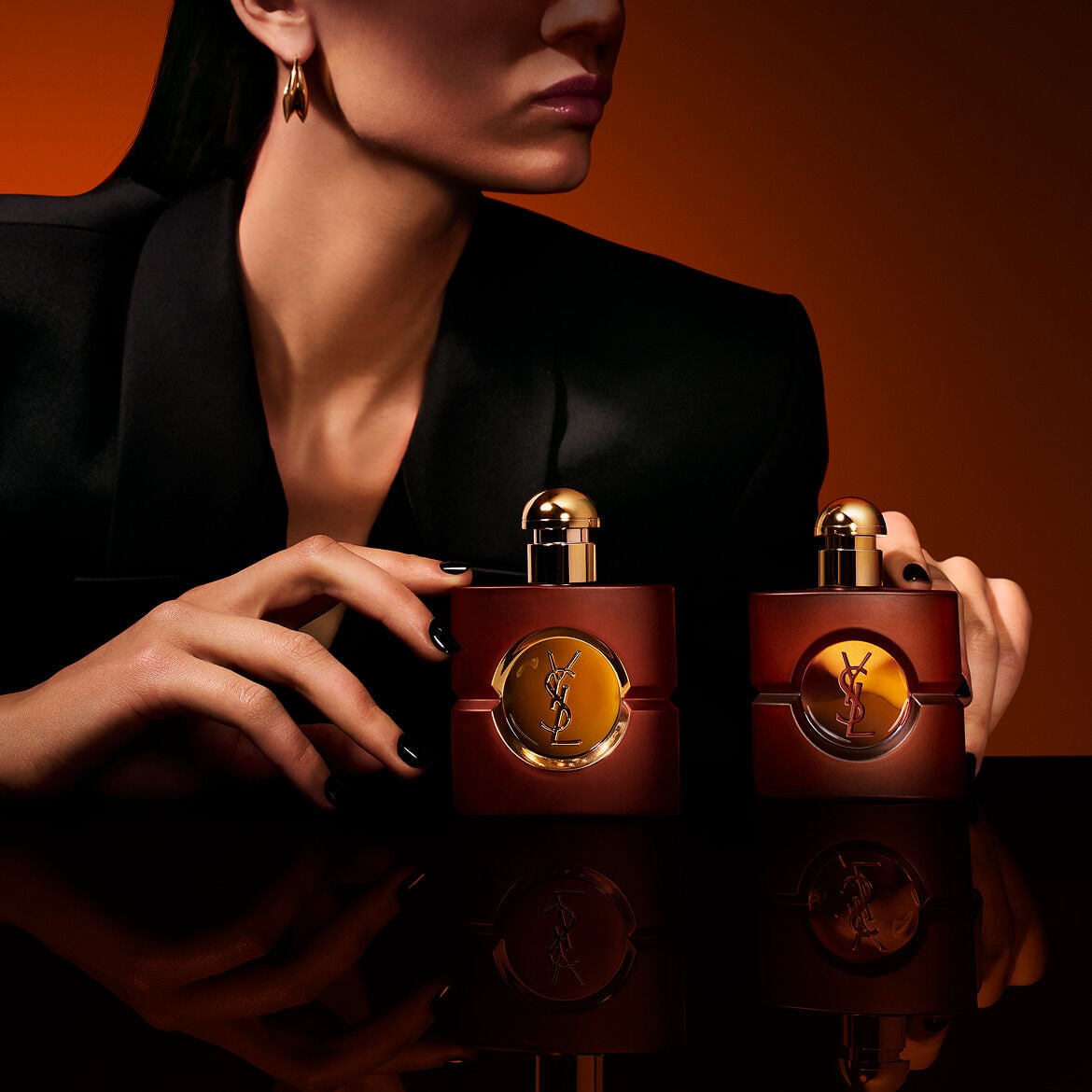 Deux flacons carrés de parfum OPIUM d&rsquo;YSL en verre dépoli, posés sur une surface noire réfléchissante, encadrés par les mains d&rsquo;une mannequin aux ongles vernis noirs, sur fond ambré chaleureux.